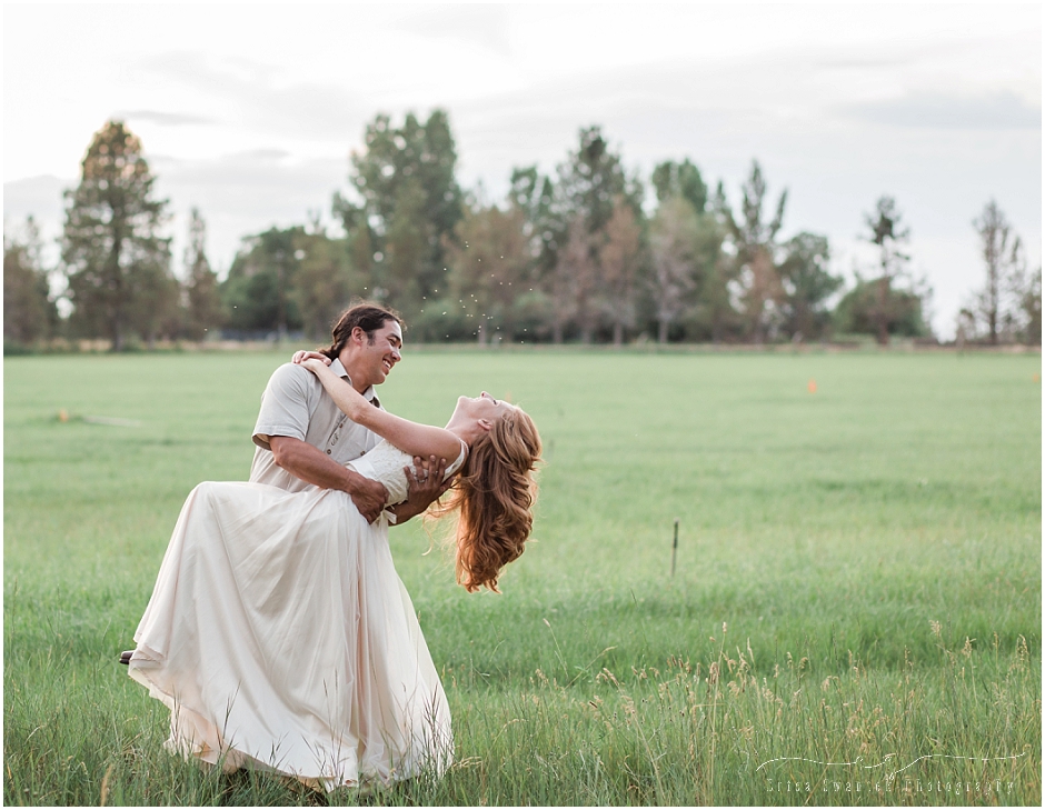 A groom wearing tan linen dips his bride wearing a blush flowing dress in a grassy field with juniper trees behind them at their rustic wedding in Bend, Oregon. | Erica Swantek Photography