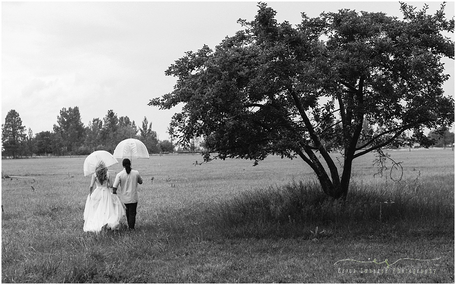 A beautiful black and white editorial style image of the bride & groom walking together away from the camera into a field holding umbrellas and hands during a rainstorm. 