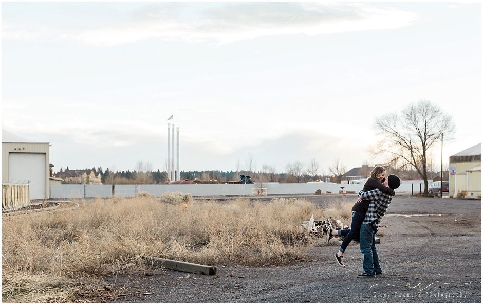A man wearing a plaid shirt and black beanie picks up a woman wearing black and kisses her with the Old Mill District smokestacks in the background of their industrial engagement photo session in Bend, Oregon. | Erica Swantek Photography