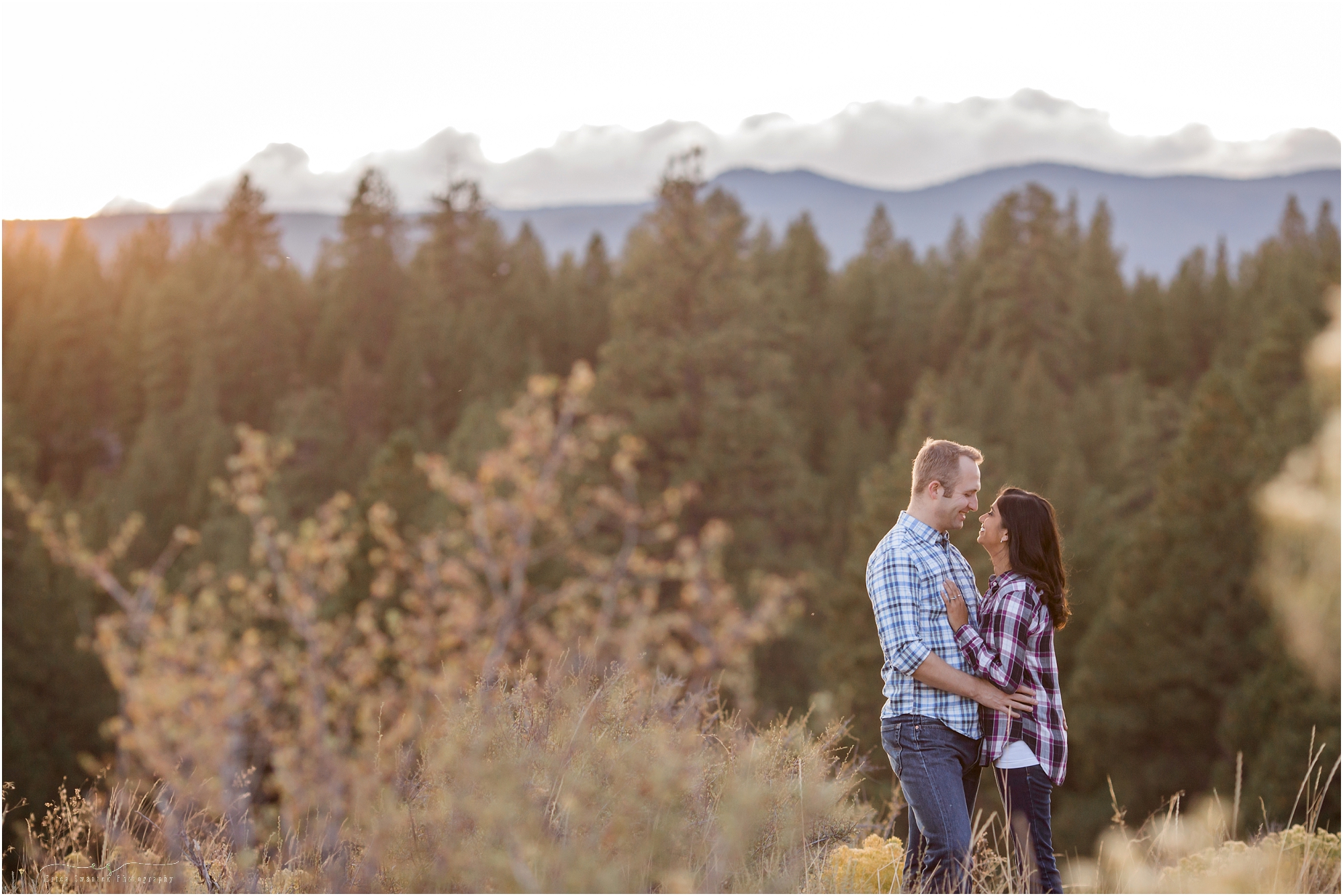 A man gently kisses a woman's forehead as they stand facing each other in their plaid flannel shirts as the sun sets behind the Oregon Cascade Mountains as viewed from the ridgeline in Bend's Shevlin Park during their fall engagement session. | Erica Swantek Photography