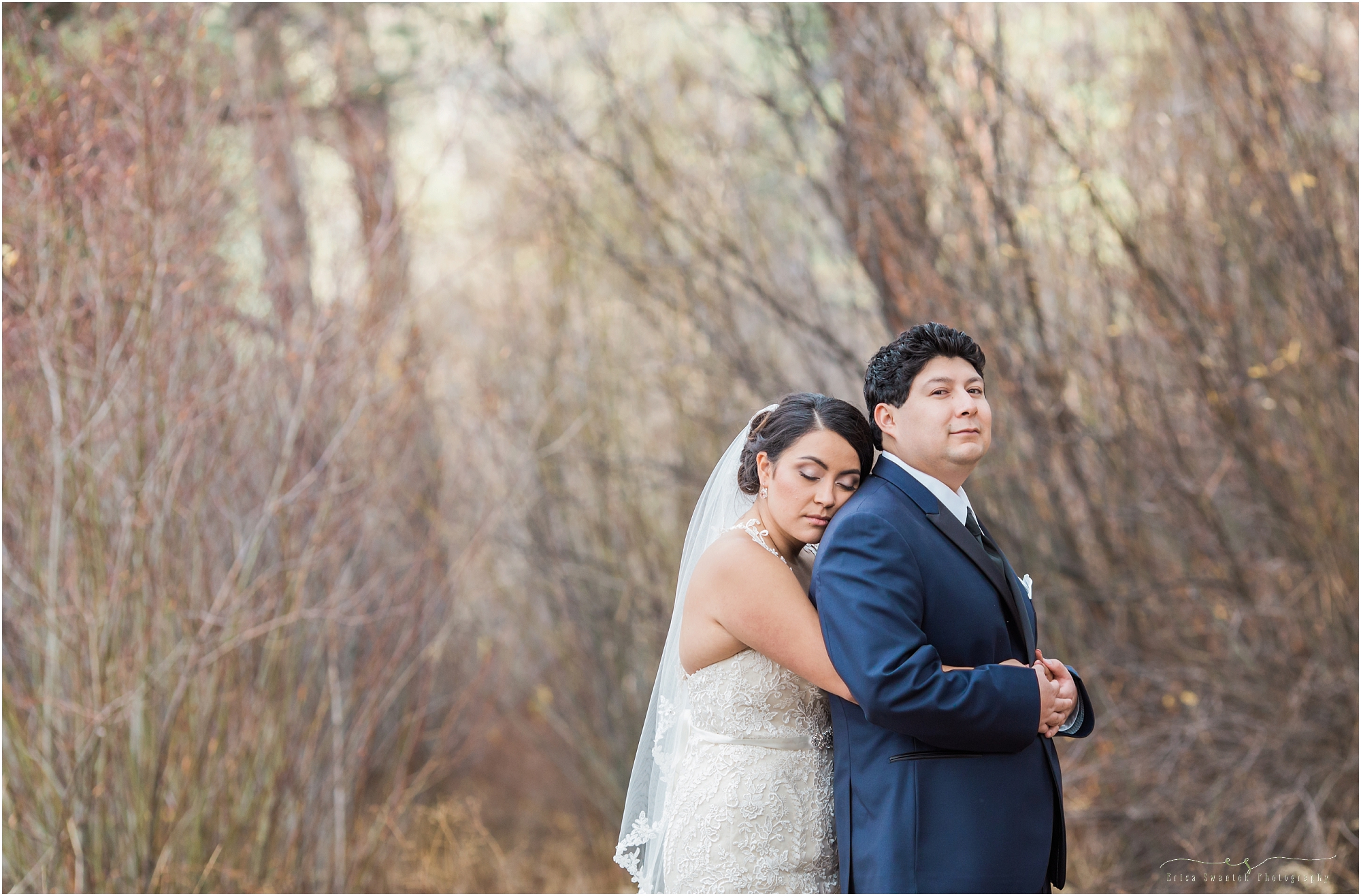 A gorgeous Mexican bride wraps her arms around her stoic groom as they pose in front of the tall pines at Shevlin Park in Bend, Oregon. 