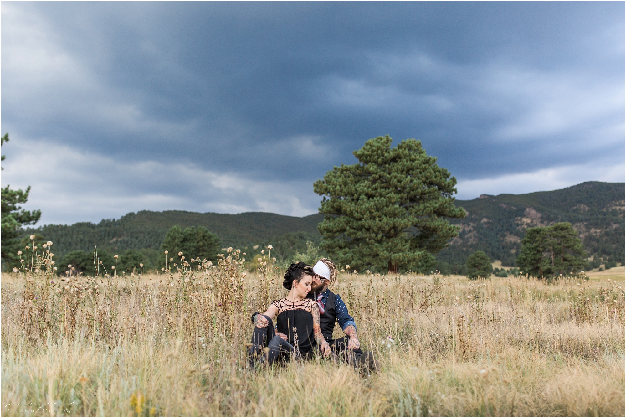 A tattooed couple sits in the tall, dry grasses of an open meadow surrounded by mountains in Colorado for their engagement session. | Erica Swantek Photography