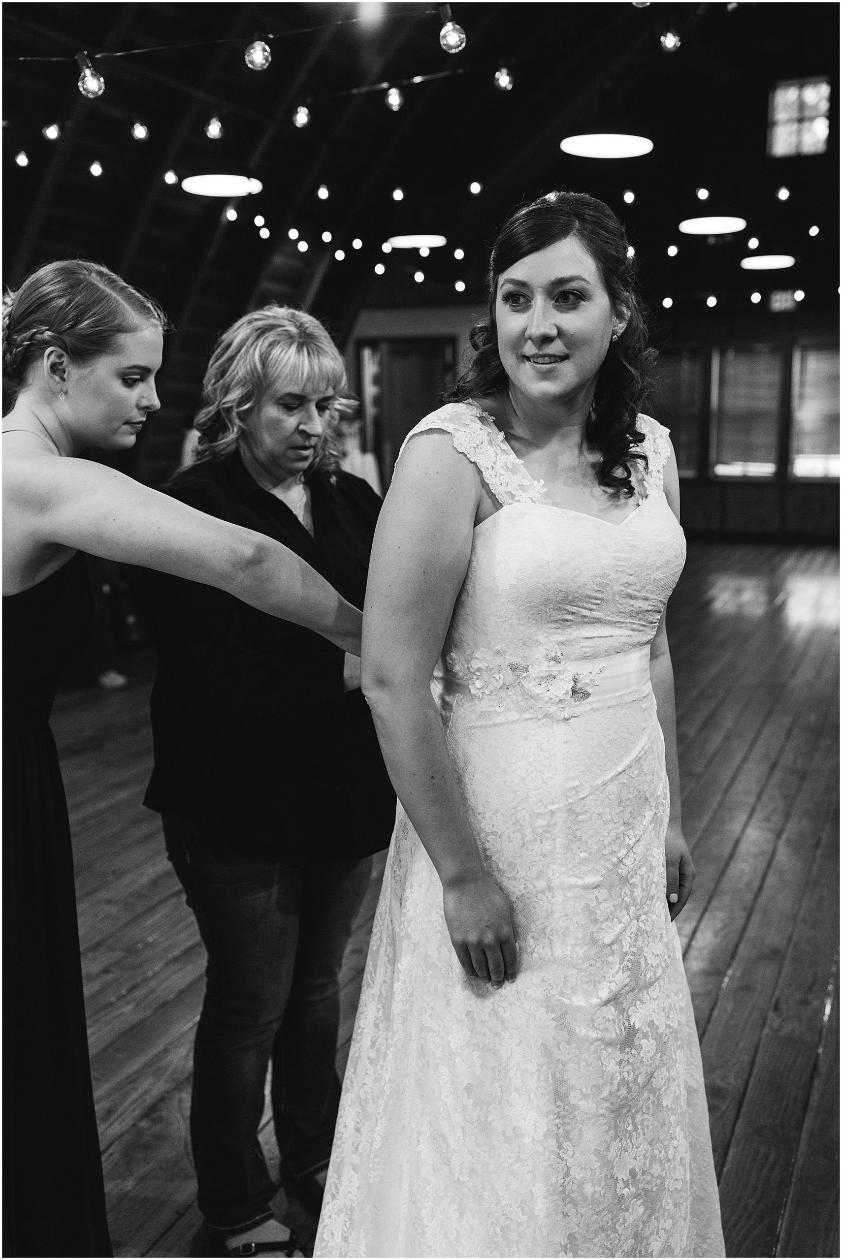 The bride's mother and maid of honor help place her sash around her waist in this beautiful black and white image from a wedding at Hollinshead Barn in Bend, Oregon. | Erica Swantek Photography