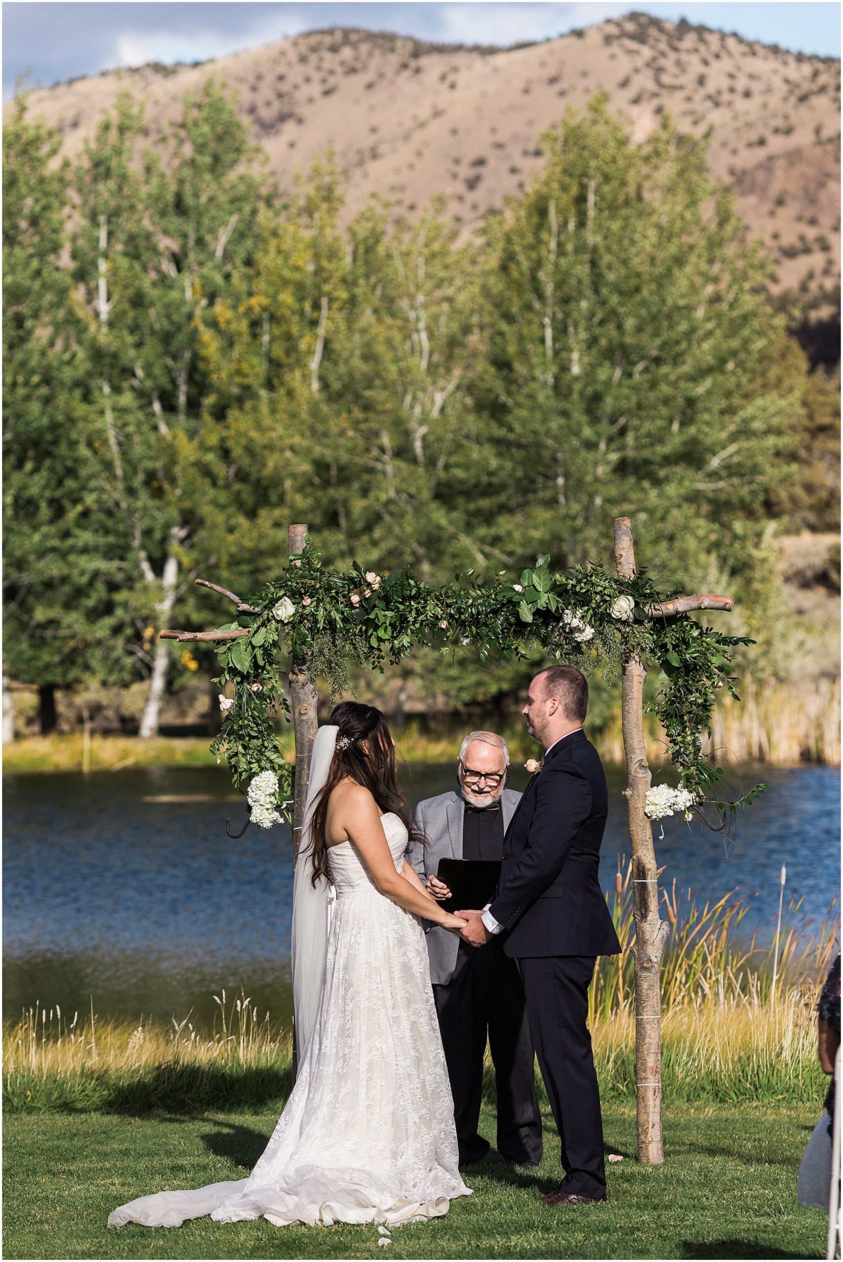 The bride and groom stand in front of their flower adorned arbor situated on the lawn in front of a beautiful pond at the Tuscan Stables wedding venue at Ranch at the Canyons in Oregon. | Erica Swantek Photography