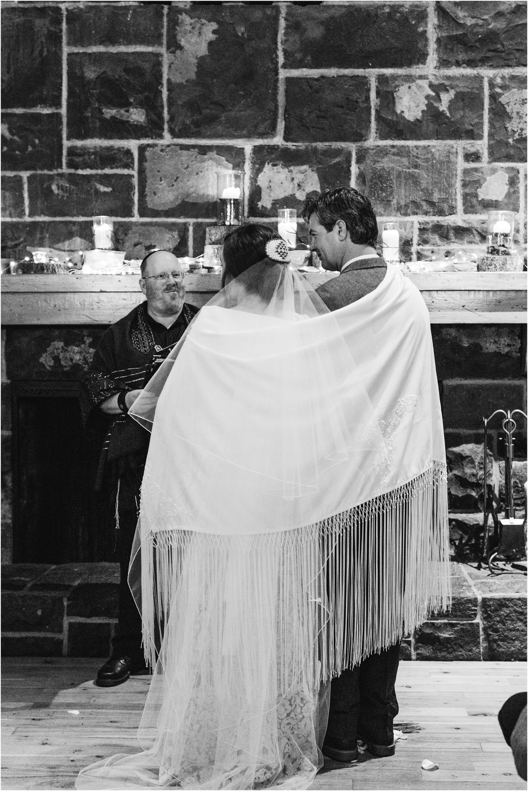 The bride and groom are wrapped in a prayer shawl incorporating Jewish and Native American traditions during their intimate ceremony at the Fireside Room at their Sunriver Resort winter wedding in Oregon. | Erica Swantek Photography