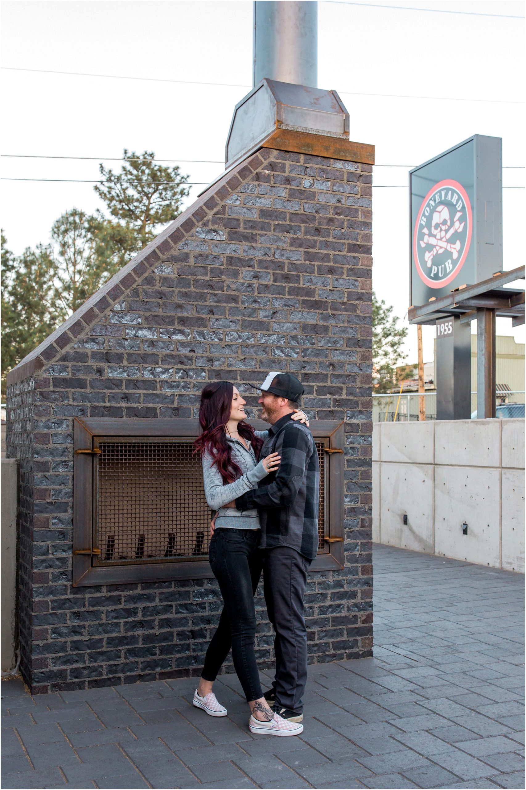 A couple poses in front of the outdoor stone fireplace at Boneyard Beer during their Bend engagement session with dogs in Oregon. | Erica Swantek Photography
