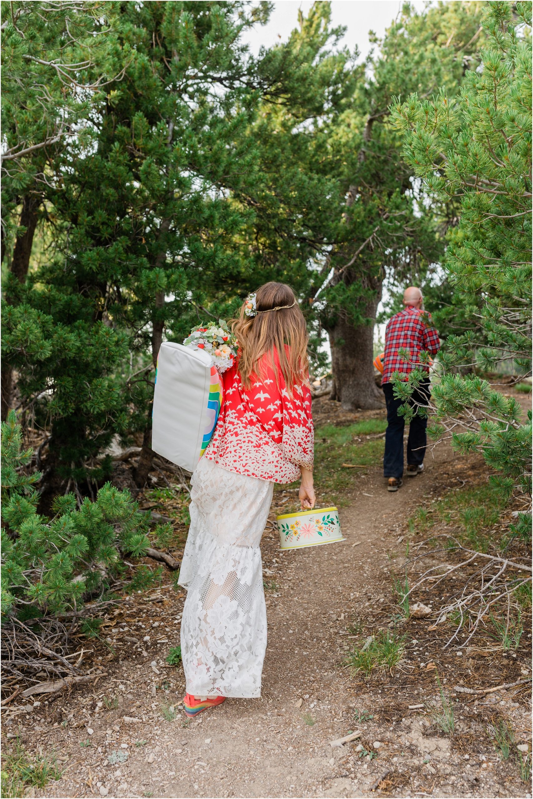 A boho bride, wearing a bright red shawl, carries a rainbow bag and vintage metal cake carrier to set up her celebratory picnic in the trees surrounding Cloud Cap Overlook for her Crater Lake Elopement in Oregon. | Erica Swantek Photography