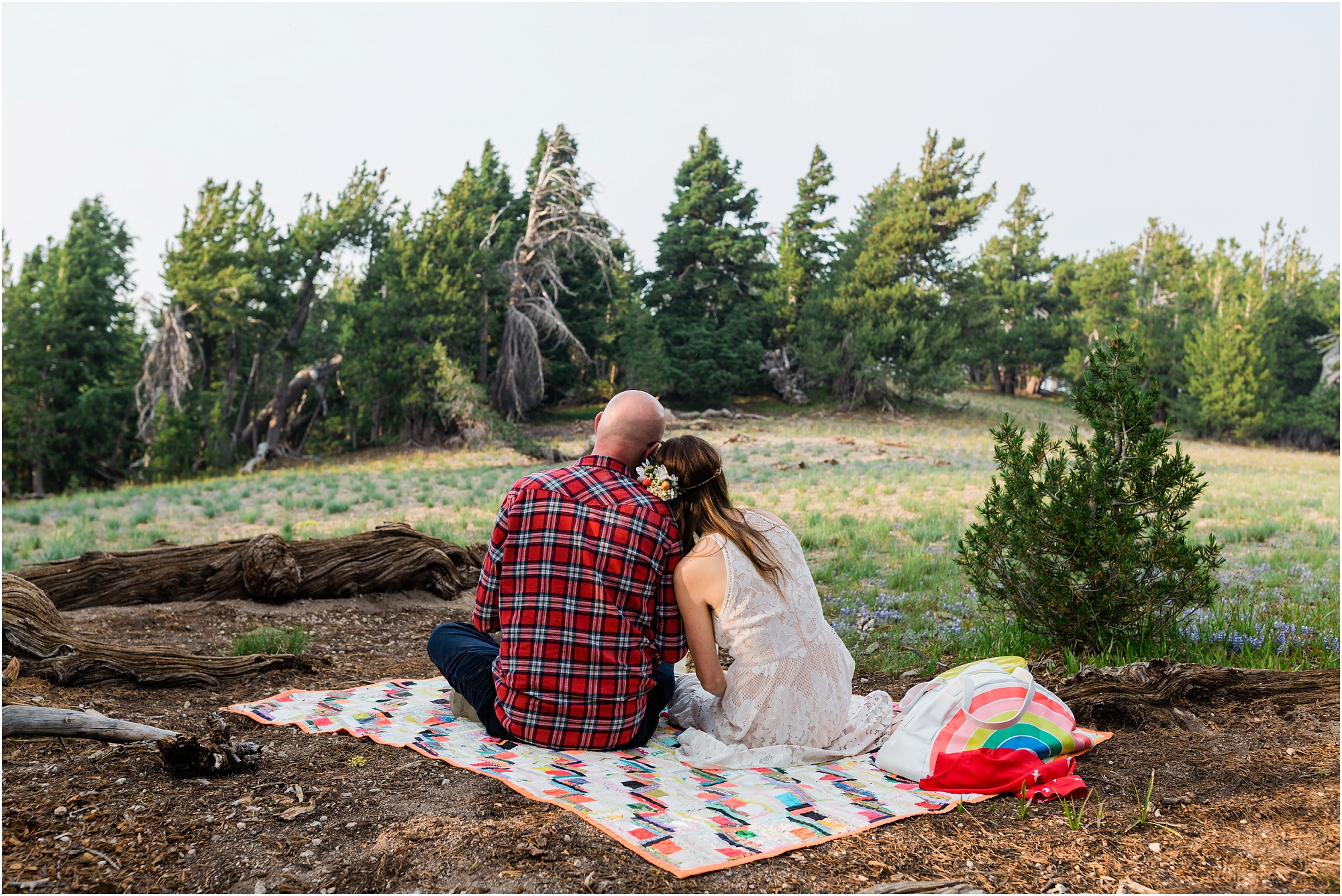 A groom wearing a red plaid shirt, and a bohemian bride, look out to the open landscape surrounding Crater Lake, after they celebrate their elopement with a toast and cake tucked into the trees near Cloud Cap Overlook at Crater Lake National Park in Oregon. | Erica Swantek Photography