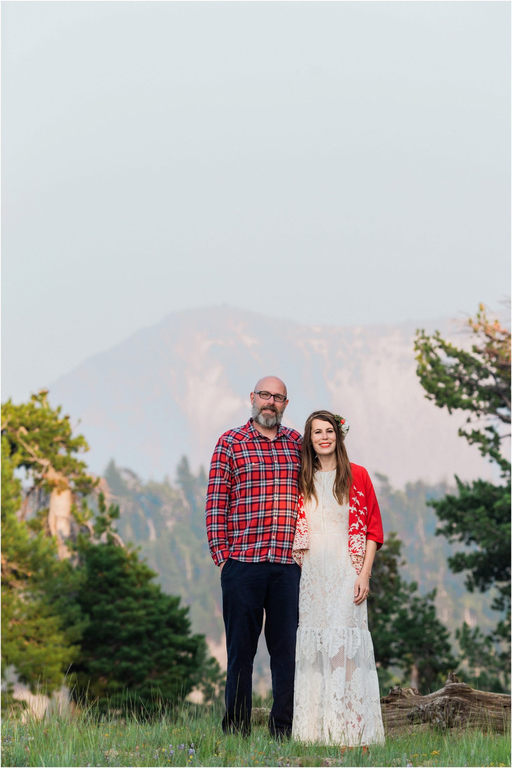 A groom wearing a red plaid shirt and navy slacks, poses with his bride, wearing a white lacy bohemian sundress, with a bright red shawl adorned with birds, pose in a meadow inside Crater Lake National Park after their elopement. | Erica Swantek Photography