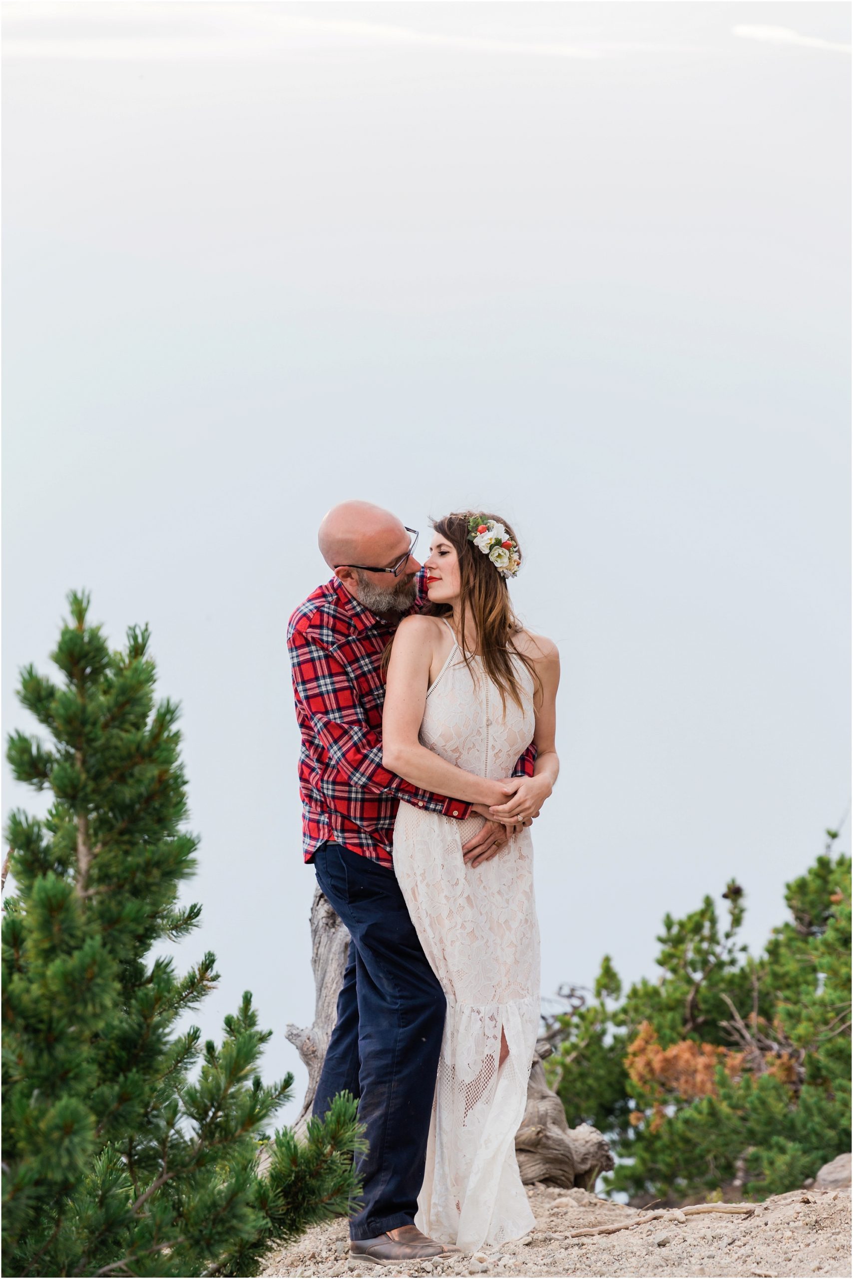 A romantic embrace as this groom wearing red plaid wraps his arms around his beautiful boho bride and goes in for a gentle kiss with Crater Lake in the background. | Erica Swantek Photography