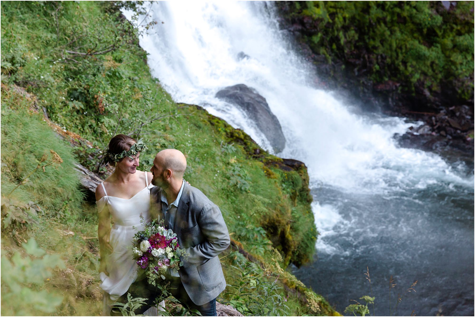 Tumalo Creek rages down the base of the waterfall as a bride wearing a floral crown looks at her groom carrying her bouquet of roses, dahlias and eucalyptus after their Bend Oregon elopement. | Erica Swantek Photography 