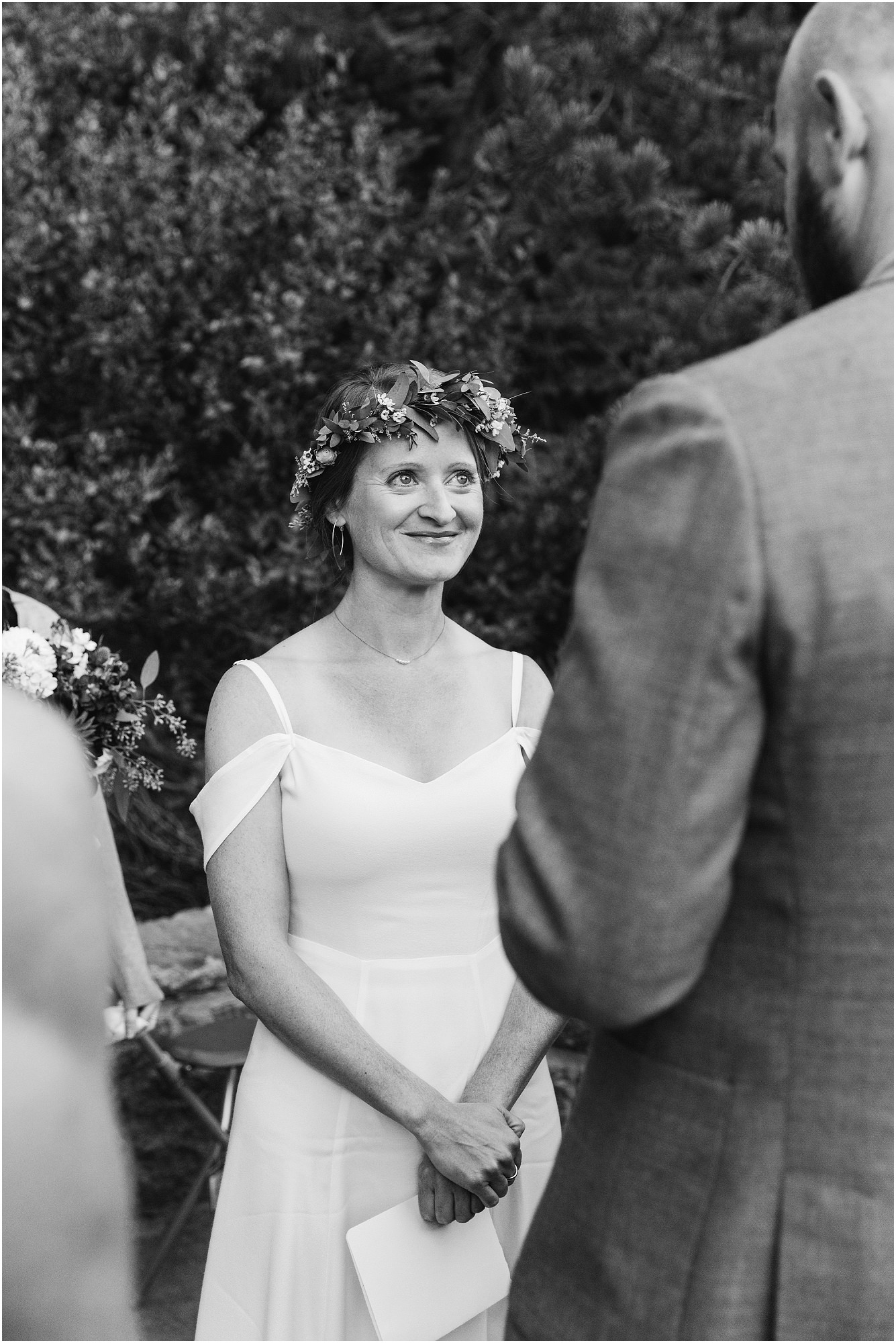 A gorgeous black and white photo of a bride wearing a floral crown smiling up at her groom as he reads his vows during their micro wedding at Tumalo Falls in Bend, Oregon. | Erica Swantek Photography