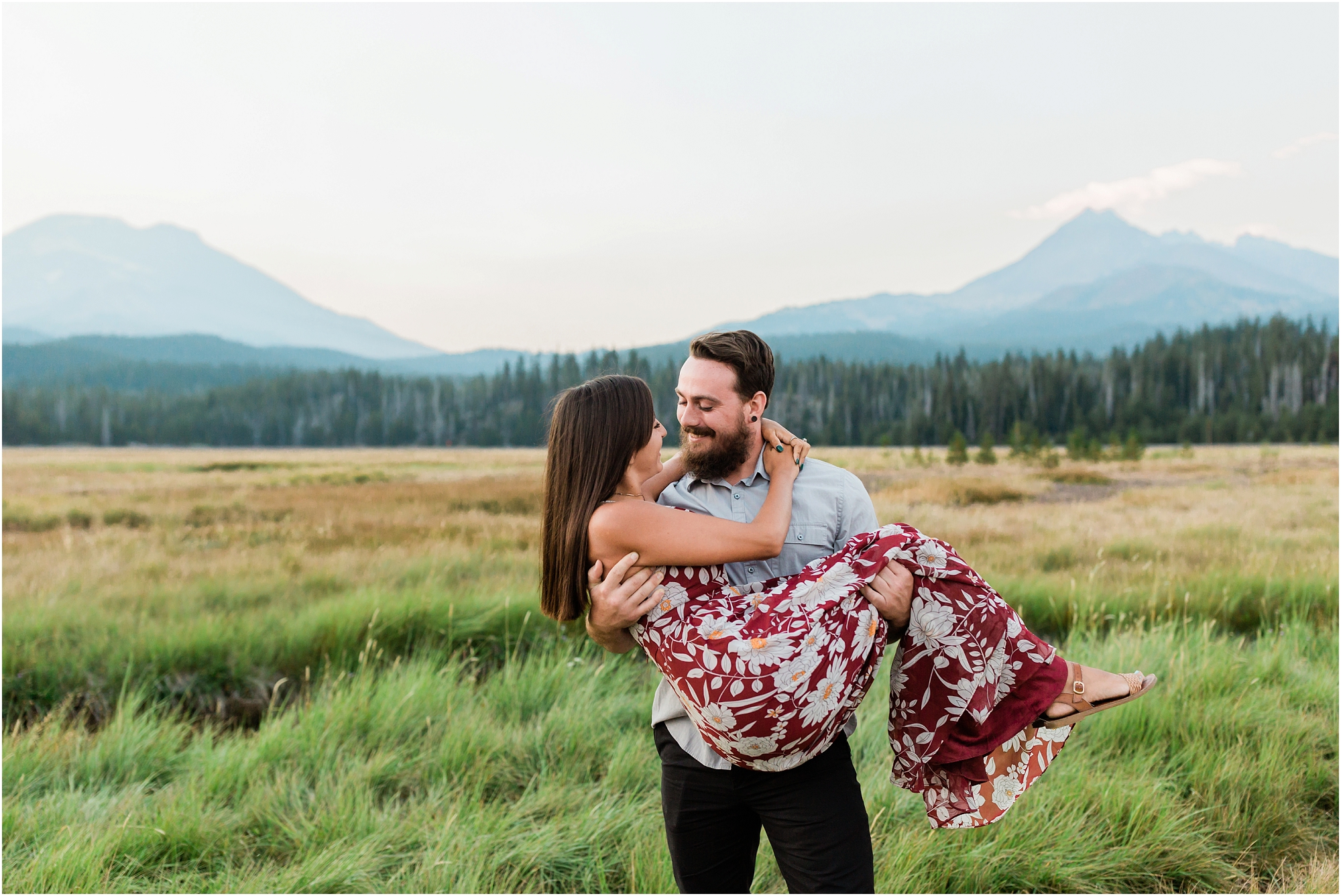 A tattooed, bearded man picks up his gorgeous woman wearing a red floral dress in a mountain meadow for their Sparks Lake Oregon engagement session. | Erica Swantek Photography