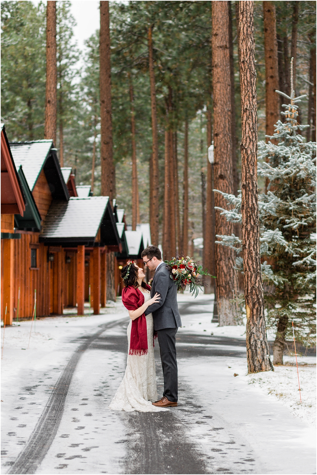 A bride, wearing a floral crown and red shawl over her white dress stands on a snowy walkway facing her groom, wearing a gray and burgundy suit, as she tosses her bouquet over his shoulder at Five Pine Lodge for their Central Oregon winter elopement. | Erica Swantek Photography 