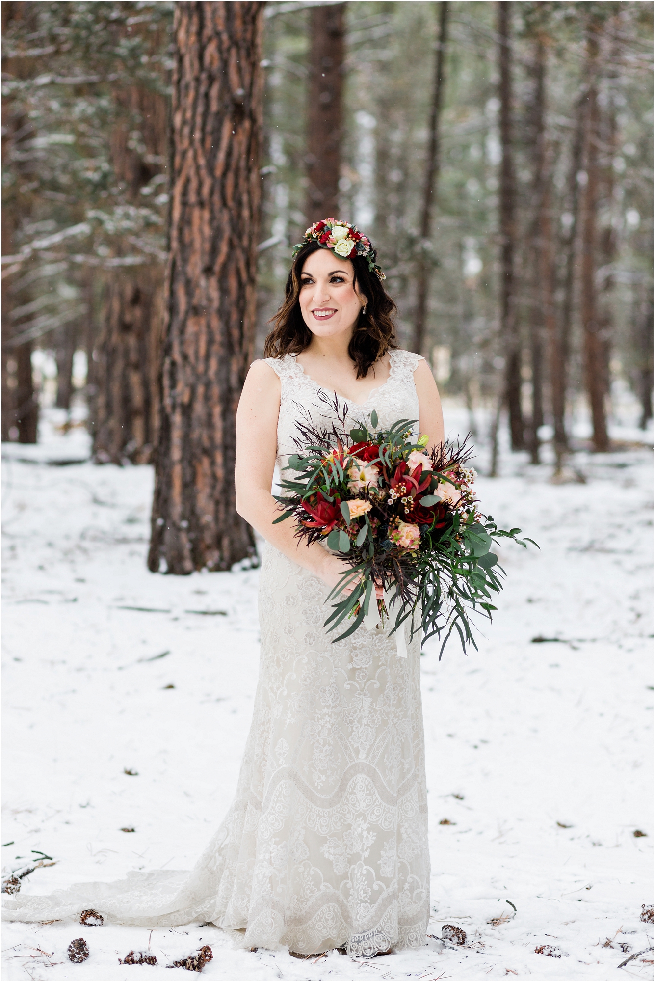 A stunning bride, wearing a white sweetheart neckline lace gown, a floral crown and gorgeous bouquet of red and pink roses and greenery sprays stands in the snow covered pines at her Five Pine Lodge Central Oregon winter elopement in Sisters. | Erica Swantek Photography
