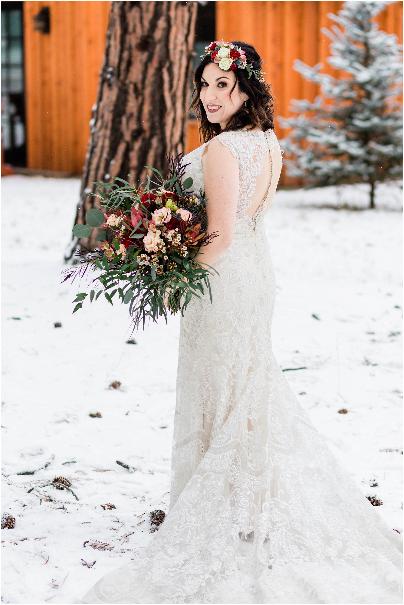 A stunning bride, wearing a white dress with an open keyhole back with buttons, a floral crown and gorgeous bouquet of red and pink roses and greenery sprays stands in the snow covered pines at her Five Pine Lodge Central Oregon winter elopement in Sisters. | Erica Swantek Photography