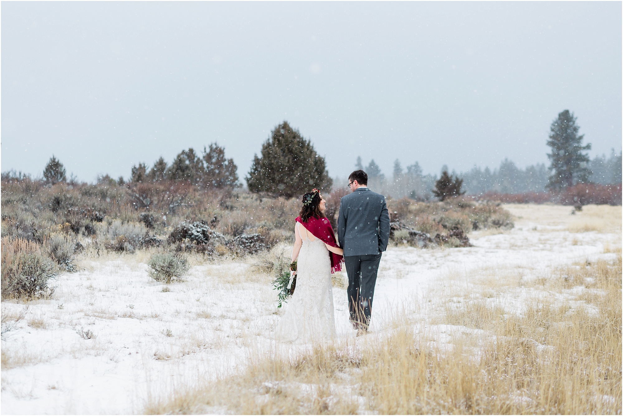 A bride wearing white with a red shawl, and a groom with a gray suit with burgundy accents walks along a snowy path at their intimate Central Oregon winter elopement near Five Pine Lodge in Sisters. | Erica Swantek Photography