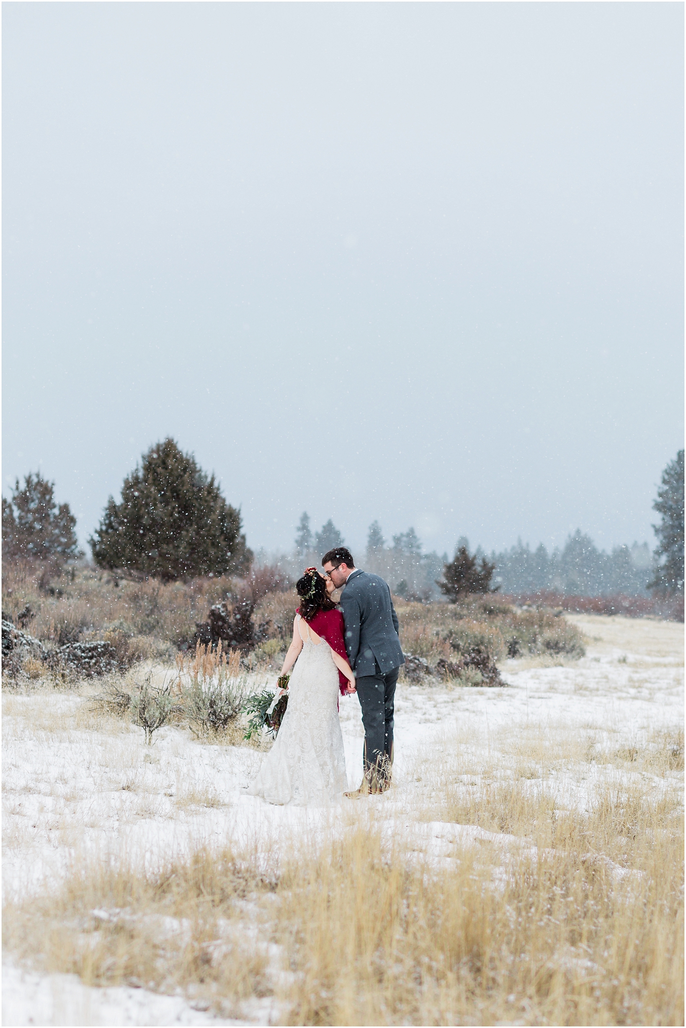A bride wearing white with a red shawl, and a groom with a gray suit with burgundy accents walks along a snowy path at their intimate Central Oregon winter elopement near Five Pine Lodge in Sisters. | Erica Swantek Photography