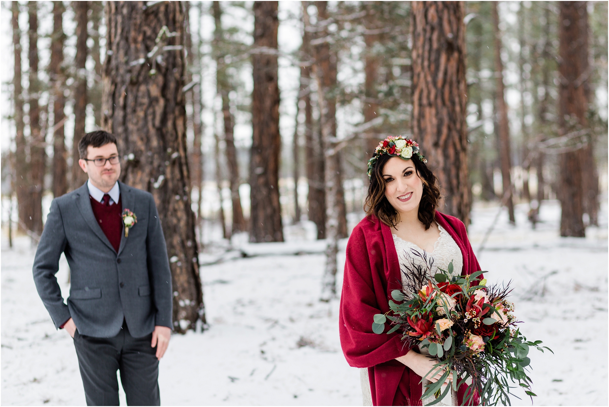 A bride, wrapped in a red shawl holding a gorgeous bouquet looks at the camera as her groom is slightly out of focus behind her in gray and burgundy, for their Central Oregon winter elopement at Five Pine Lodge in Sisters. | Erica Swantek Photography