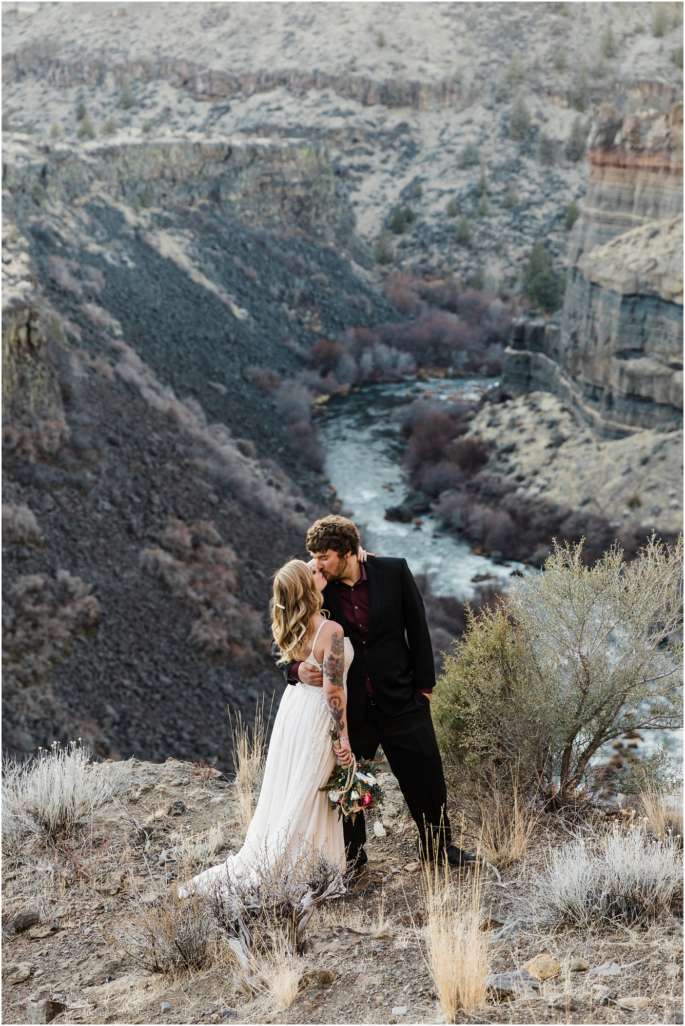 A tattooed boho bride, wearing a lace dress, kisses her groom in black and burgundy overlooking the Deschutes River for an amazing adventure elopement near Bend Oregon. | Erica Swantek Photography