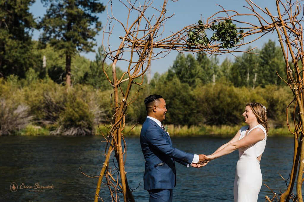 bride and groom holding hands under a wooden arch by a river shore