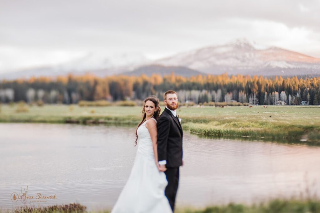 bride and groom holding hands next to a large lake with mountain views at the black butte ranch