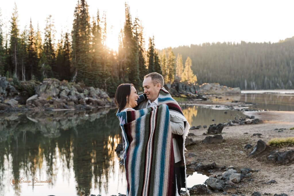 elopement couple standing wrapped in a blanket by a river shore with mountain views in the background