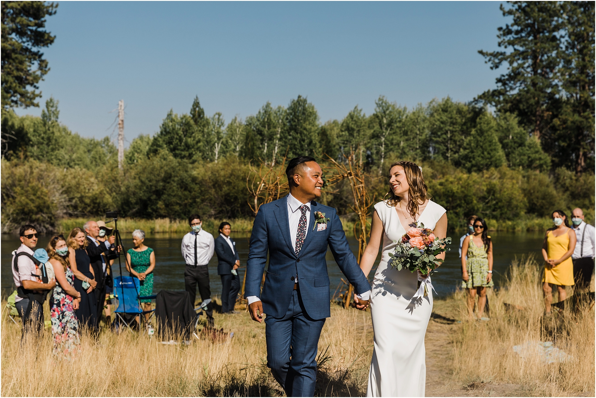 A newly married couple walks hand in hand as their wedding guests stand behind them on the edge of the river bank after their small elopement ceremony in Bend, Oregon. | Erica Swantek Photography