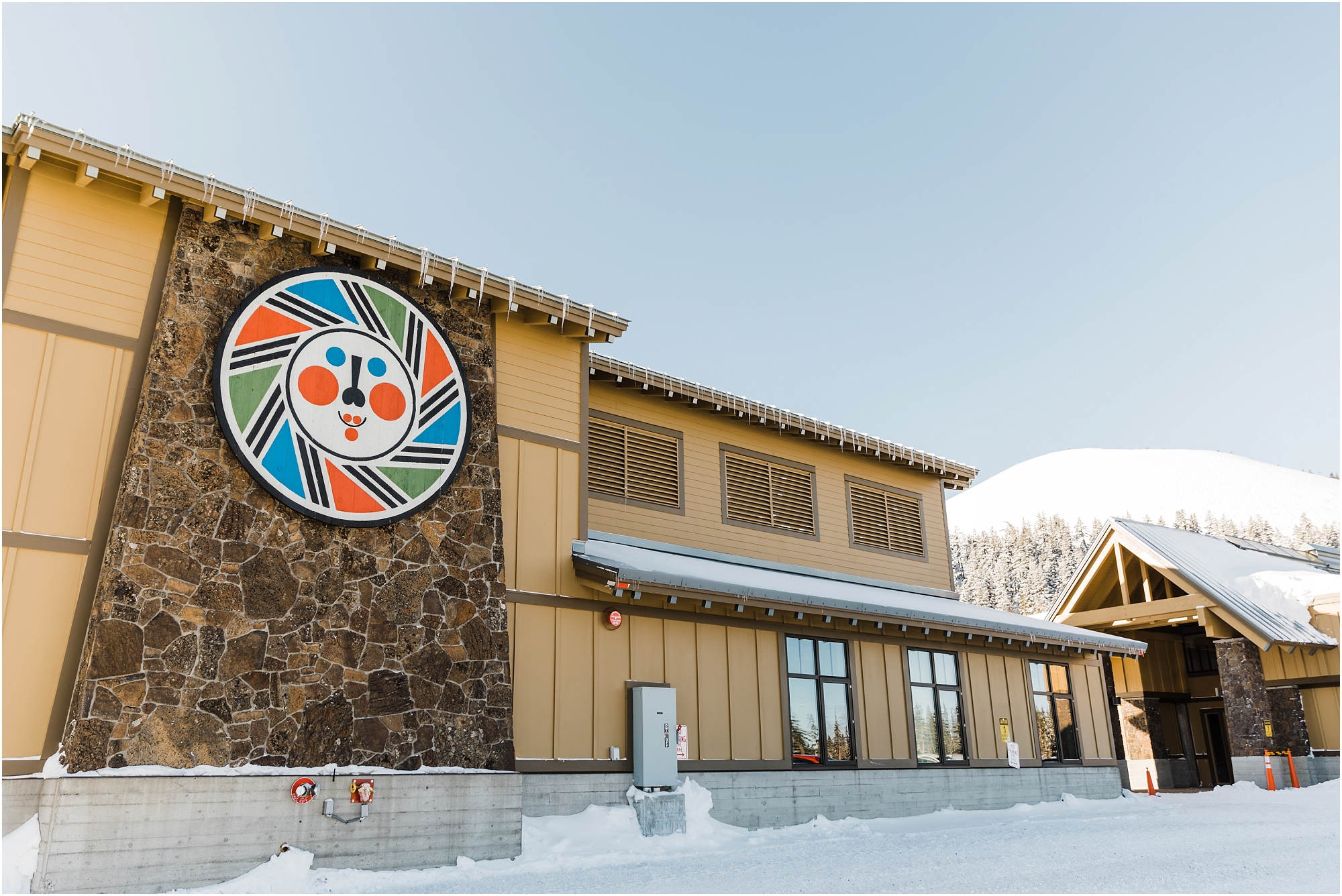 A two story building with the Mt Bachelor logo greets guests in the snow covered parking lot as the snow covered cinder butte known as the cone stands tall in the background of Mt Bachelor Ski Resort in Bend, Oregon. | Erica Swantek Photography