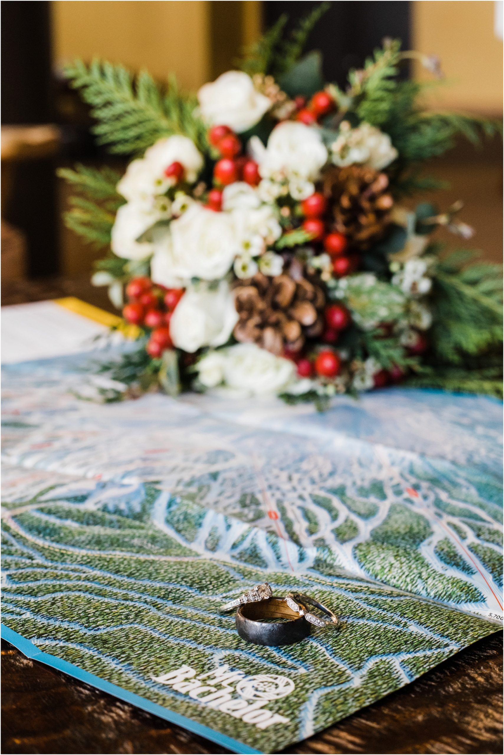 Wedding rings sit atop the Mt Bachelor trail map with a bouquet of white flowers, red berries, pine cones and green pine boughs for this winter skiing Mt Bachelor Adventure Elopement in Bend, OR. | Erica Swantek Photography