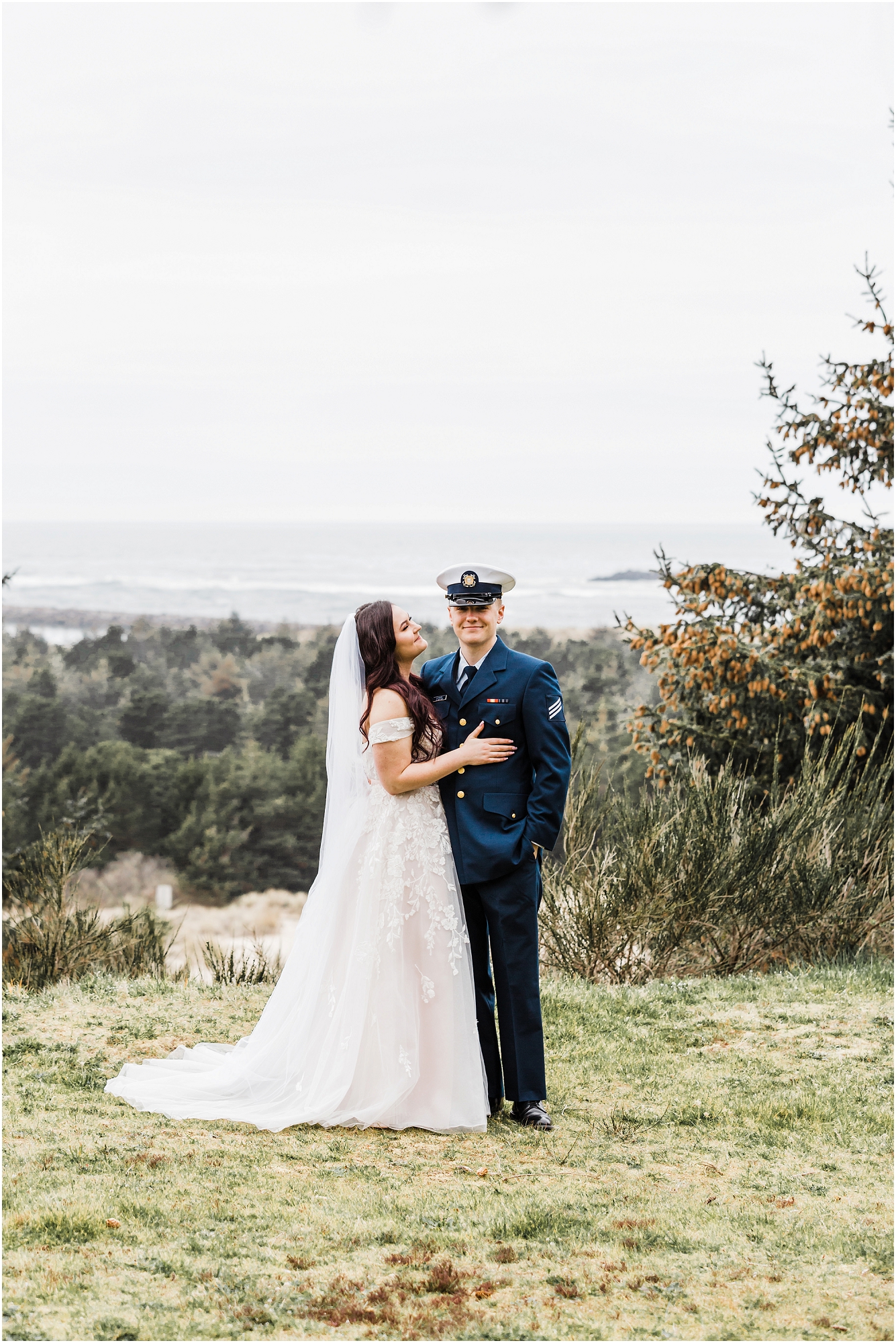 A groom, wearing his navy US Coast Guard uniform, and a bride wearing a blush pink off the shoulder gown, with a long white veil, pose together on a grassy overlook above the ocean for this Oregon Coast Guard Elopement in Winchester Bay. | Erica Swantek Photography