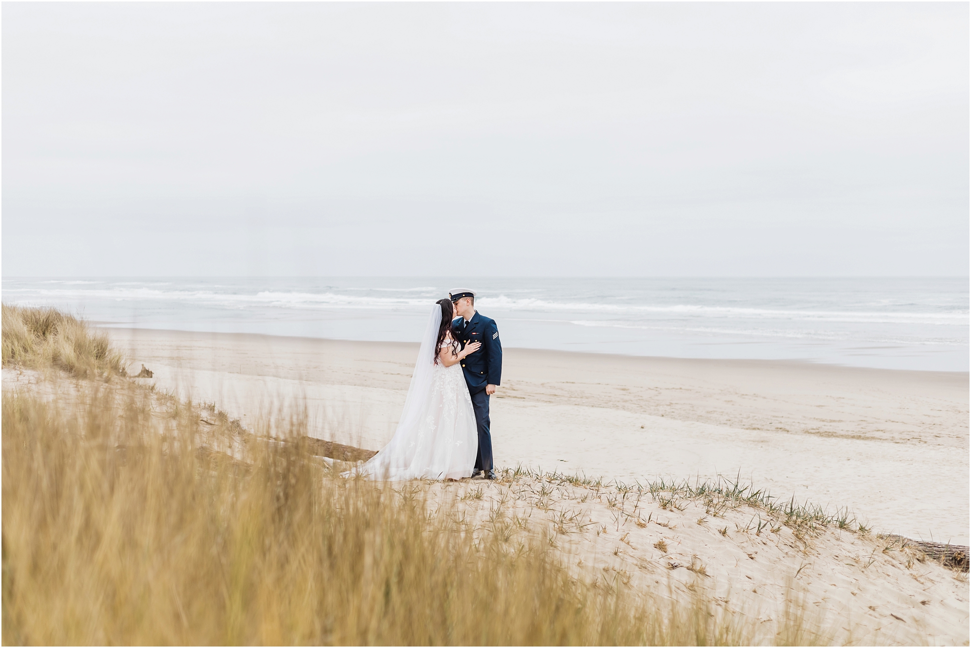 A groom, wearing his navy US Coast Guard uniform, and a bride wearing a blush pink off the shoulder gown, with a long white veil, pose together on the sandy beach along the ocean for this Oregon Coast Guard Elopement in Winchester Bay. | Erica Swantek Photography