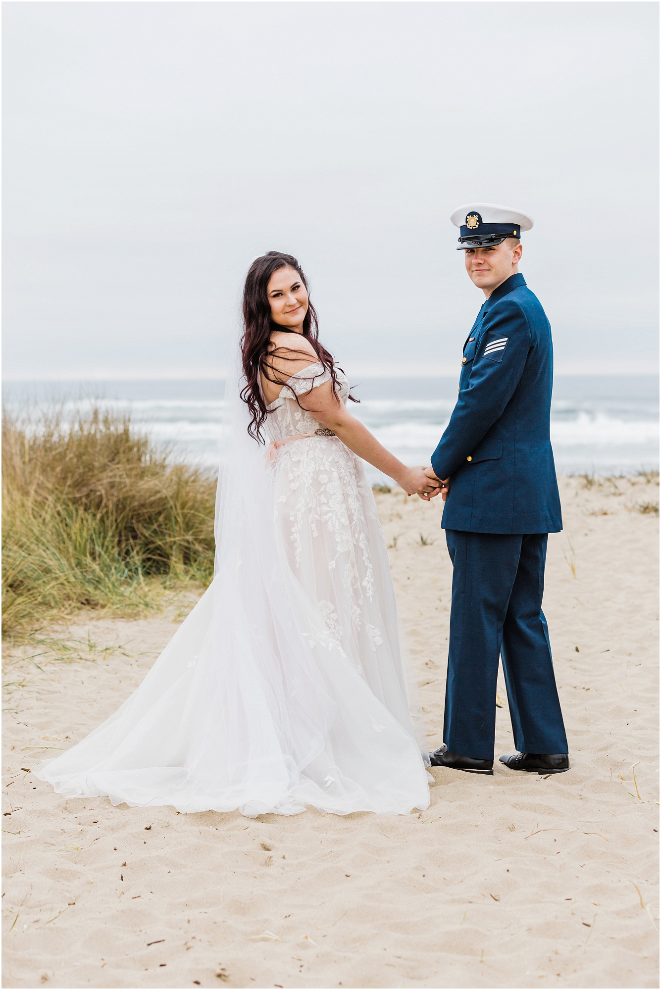 A groom, wearing his navy US Coast Guard uniform, and a bride wearing a blush pink off the shoulder gown, with a long white veil, pose together on the sandy beach along the ocean for this Oregon Coast Guard Elopement in Winchester Bay. | Erica Swantek Photography