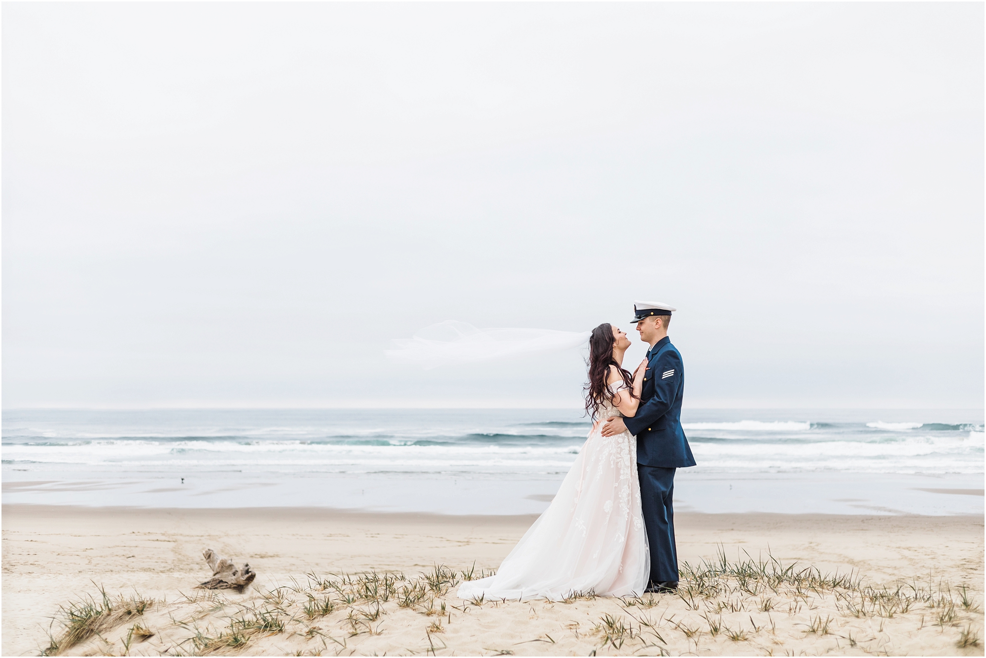 A groom, wearing his navy US Coast Guard uniform, and a bride wearing a blush pink off the shoulder gown, with a long white veil, pose together on the sandy beach along the ocean for this Oregon Coast Guard Elopement in Winchester Bay. | Erica Swantek Photography