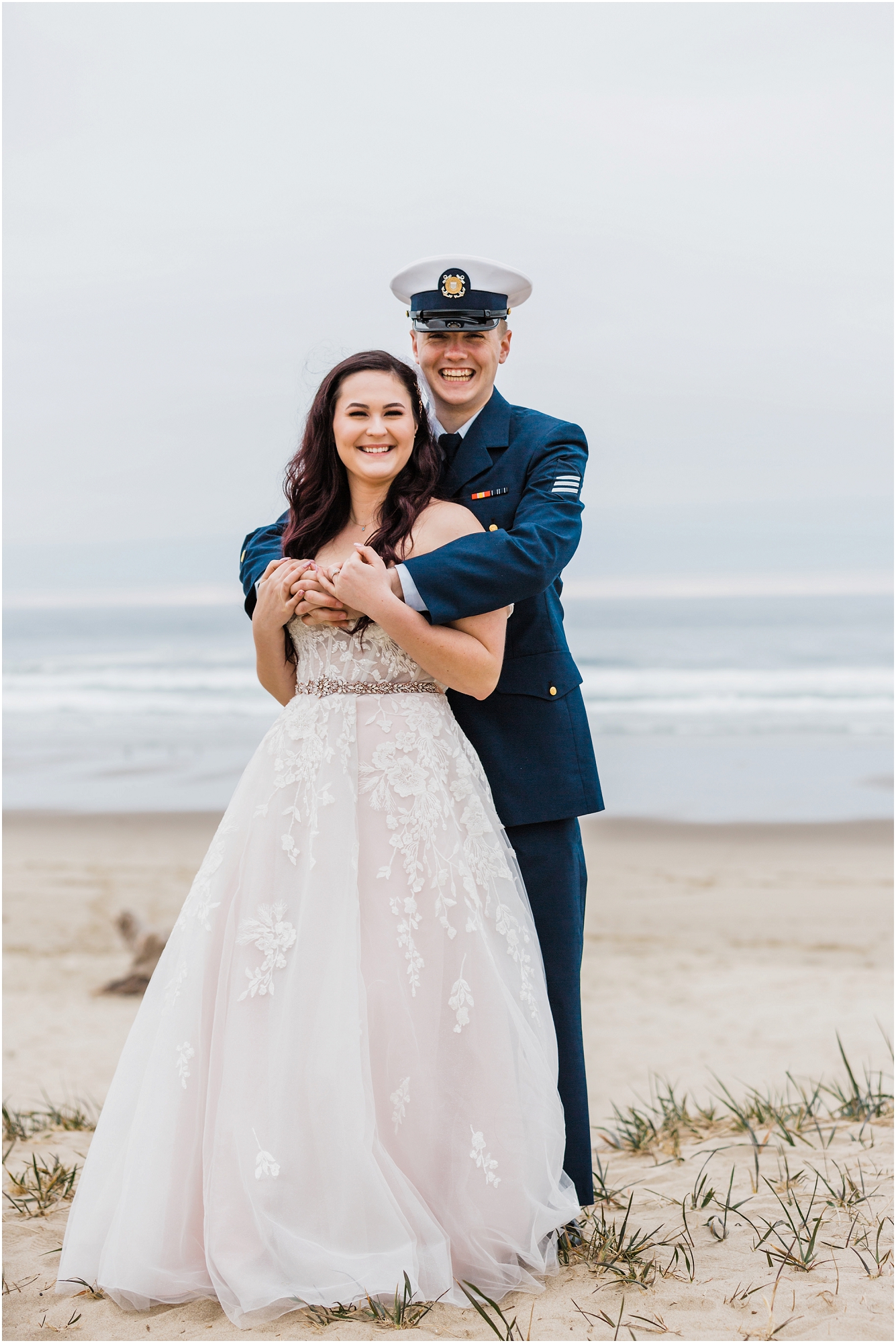 A groom, wearing his navy US Coast Guard uniform, and a bride wearing a blush pink off the shoulder gown, with a long white veil, pose together on the sandy beach along the ocean for this Oregon Coast Guard Elopement in Winchester Bay. | Erica Swantek Photography