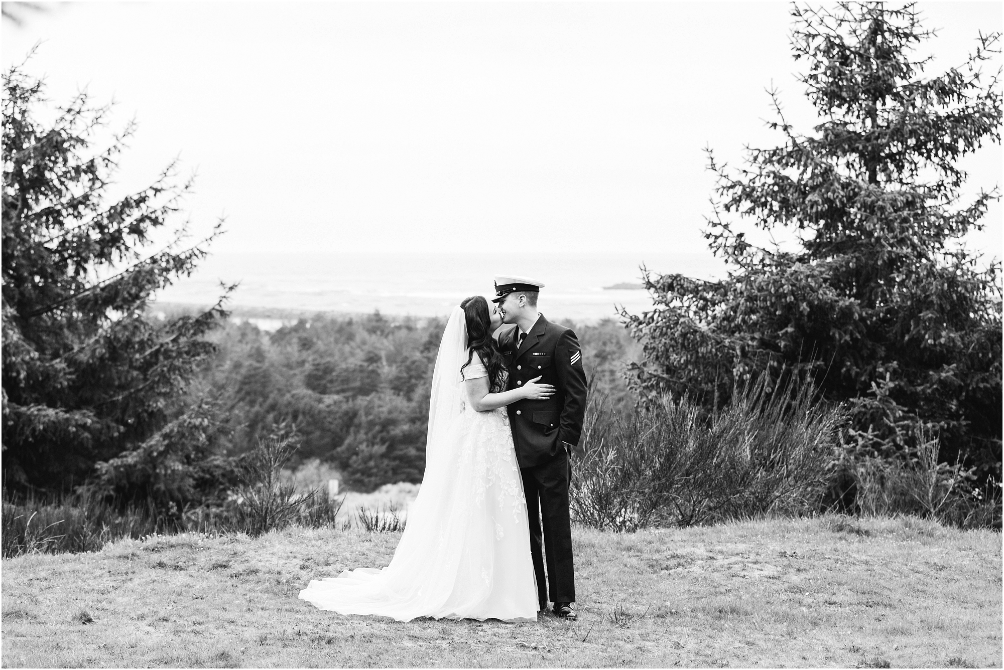 A black and white image of a groom, wearing his navy US Coast Guard uniform, and a bride wearing a blush pink off the shoulder gown, with a long white veil, pose together on a grassy overlook above the ocean for this Oregon Coast Guard Elopement in Winchester Bay. | Erica Swantek Photography