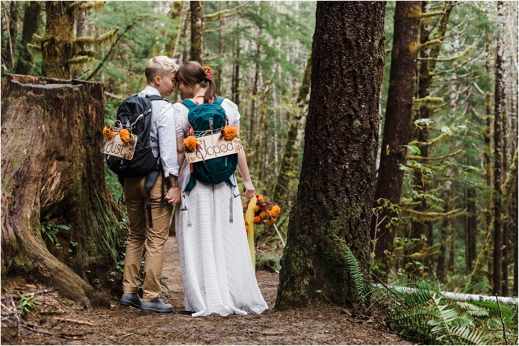 A couple stands forehead to forehead with their backs to the camera wearing backpacks decorated with Just Eloped signs on their PNW hiking adventure elopement day in Oregon. | Erica Swantek Photography