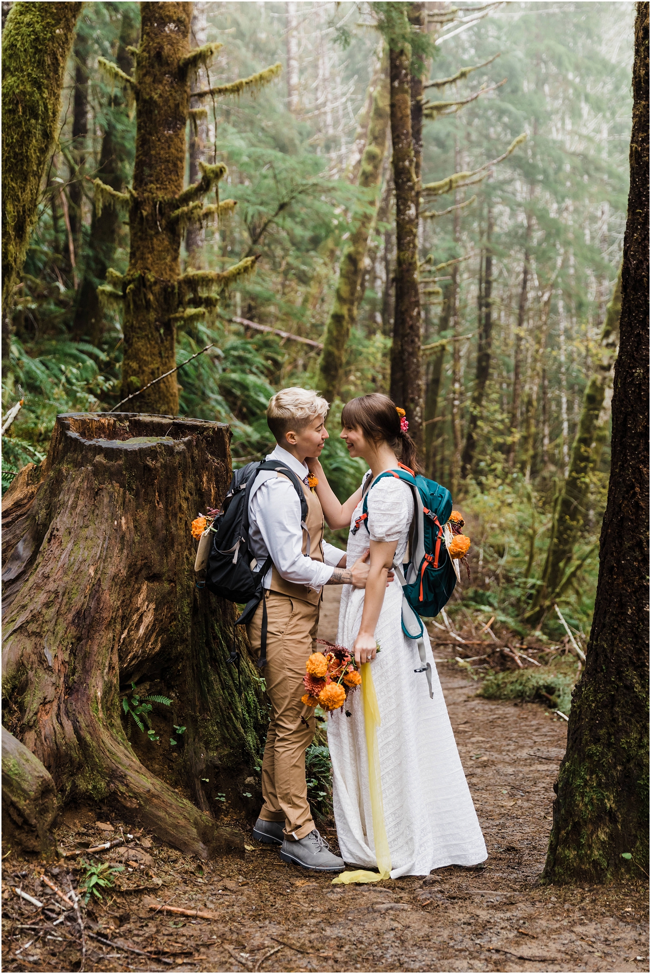 A couple wearing wedding clothing and backpacks decorated with Just Eloped signs embrace near an old growth stump as they hike to their waterfall elopement in the PNW. | Erica Swantek Photography