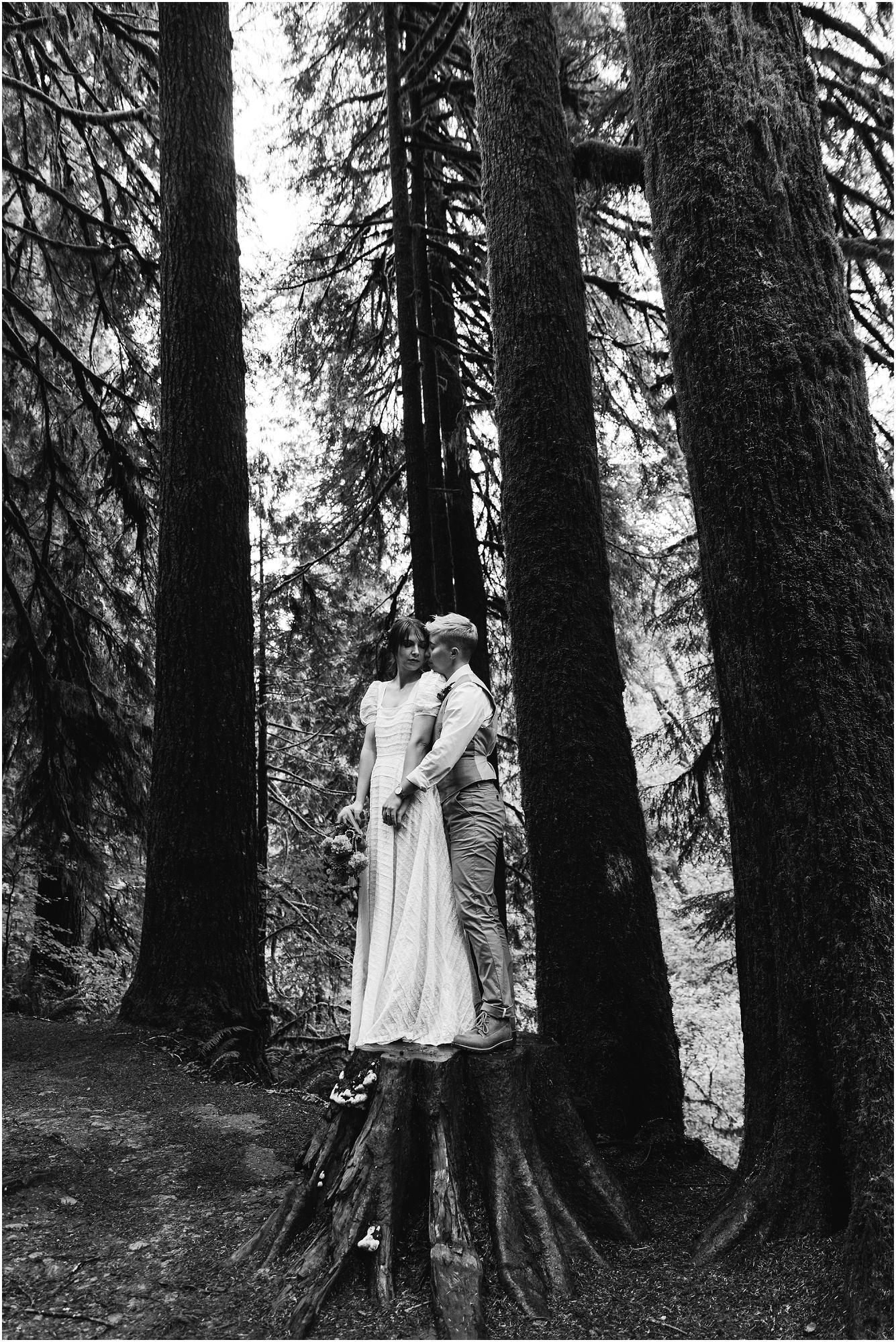 A gorgeous black and white image of a couple standing on top of a large tree stump in the forest wearing their wedding clothes and embracing their PNW hiking adventure elopement in Oregon. | Erica Swantek Photography