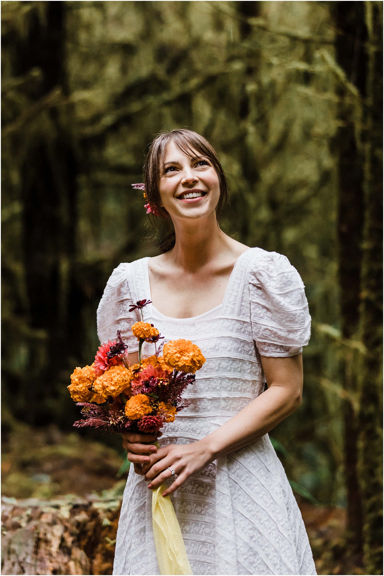 A gorgeous bride wearing a white puff sleeved BHLDN dress with an orange marigold bouquet standing in a beam of diffuse light in the dark PNW forest for her adventure elopement in Oregon. | Erica Swantek Photography