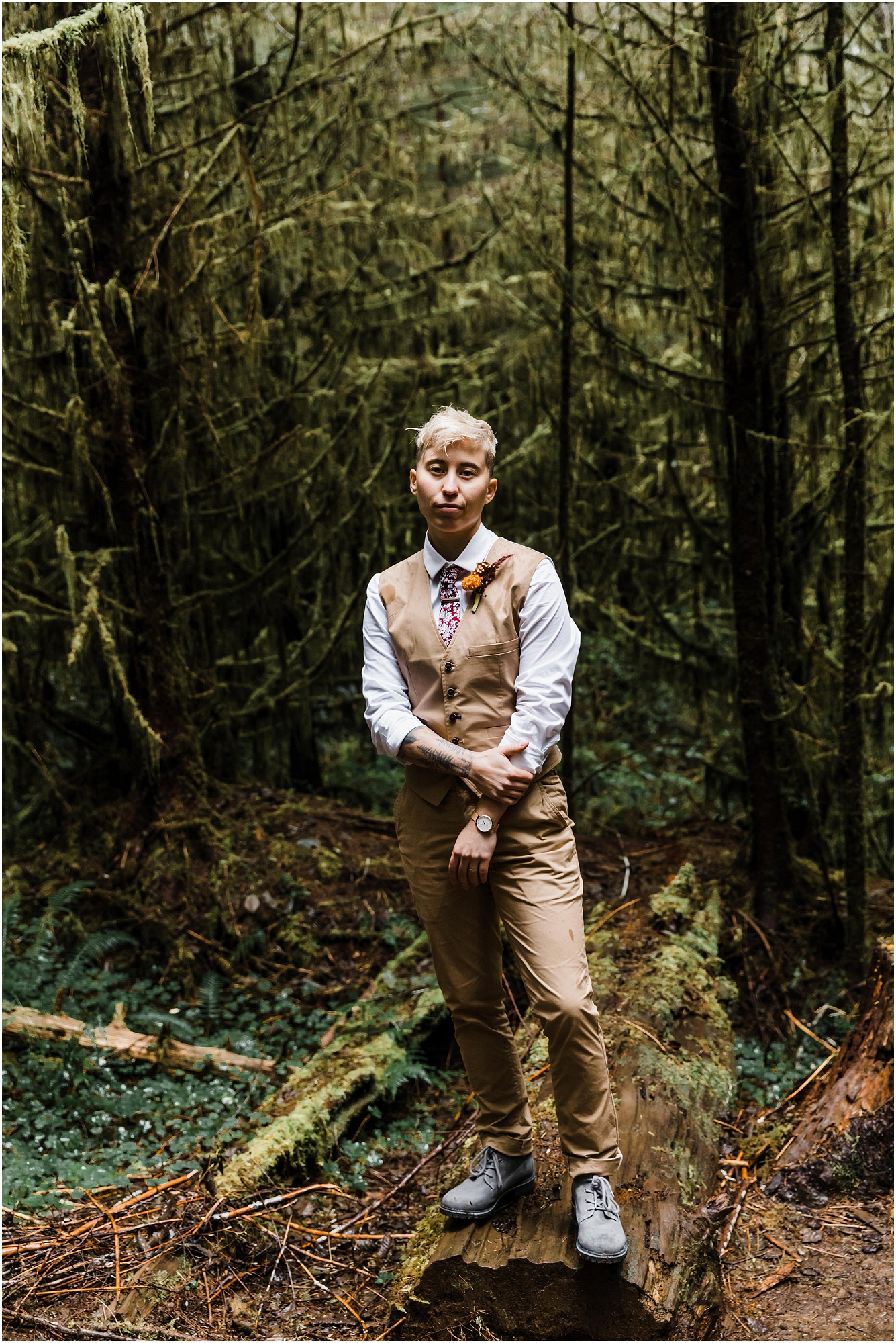 A handsome LGBTQ+ groom wearing a tan vest and pants with a light gray button down top and an orange marigold boutonniere stands among the lush mossy forest for his PNW hiking adventure elopement. | Erica Swantek Photography 
