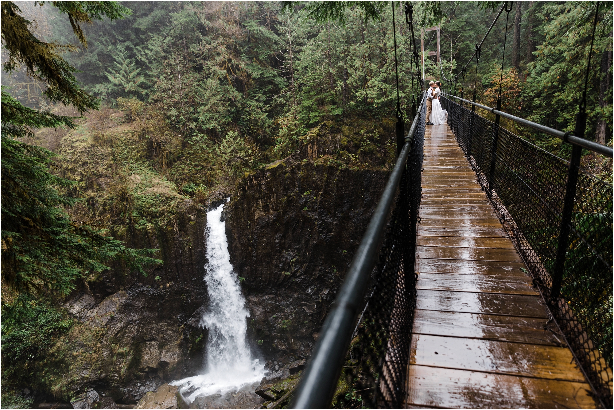 A stunning landscape of a wedding couple standing on the suspension bridge over Drift Creek falls snuggling under their wool blanket as the rain pours down on them during their PNW hiking adventure elopement. | Erica Swantek Photography