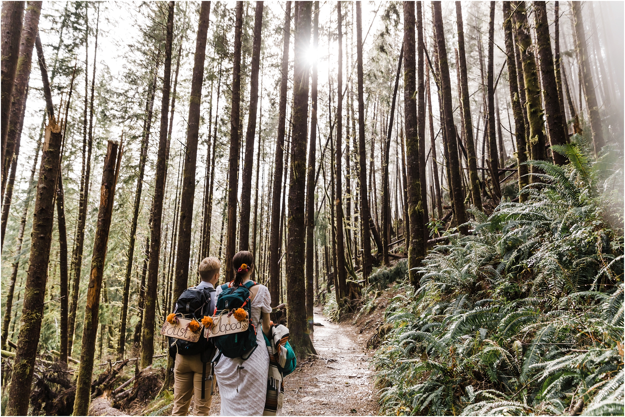 A LGBTQ+ couple hikes in the rain along a forested trail wearing backpacks with Just Eloped wooden signs during their PNW hiking adventure elopement in Oregon. | Erica Swantek Photography
