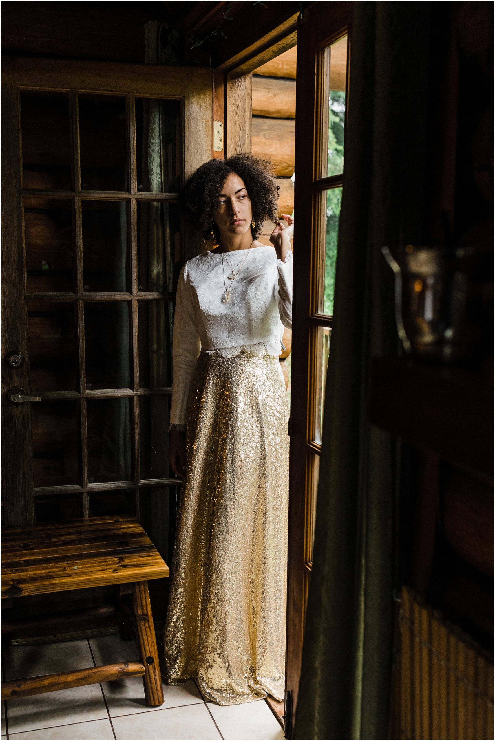 A black bride wearing a white cropped top and gold sequined skirt stands in the doorway of a rustic cabin as she gets ready for her Oregon Coast elopement on the beach at Proposal Rock in Neskowin, Oregon. | Erica Swantek Photography