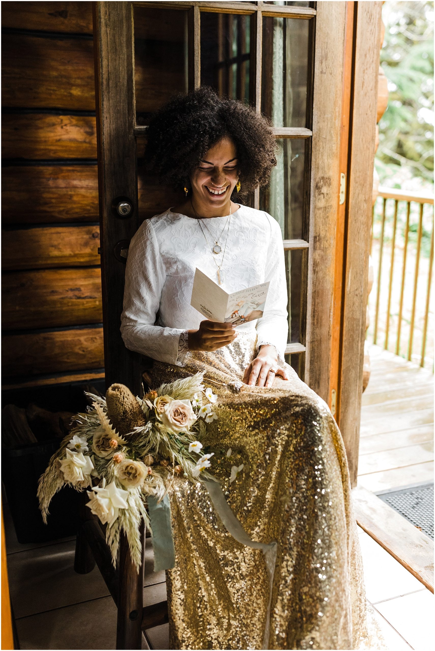 A black bride wearing a white cropped top and gold sequined skirt sits in the doorway of a rustic cabin reading a note from her fiancée as she gets ready for her Oregon Coast elopement on the beach at Proposal Rock near Lincoln City. | Erica Swantek Photography