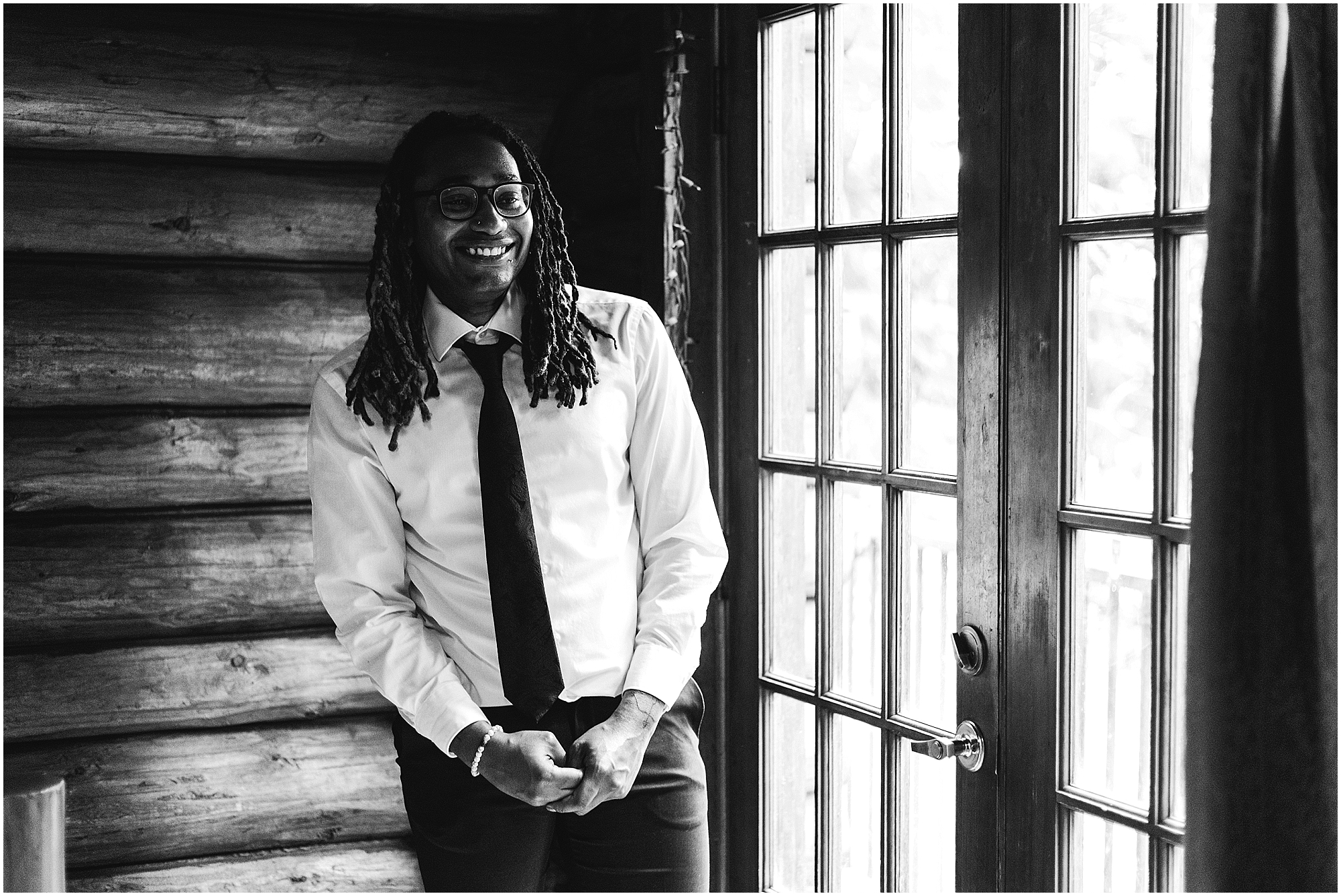 A black and white image of a black groom nervously laughing in front of french doors in a cabin the woods along the Oregon Coast. | Erica Swantek Photography