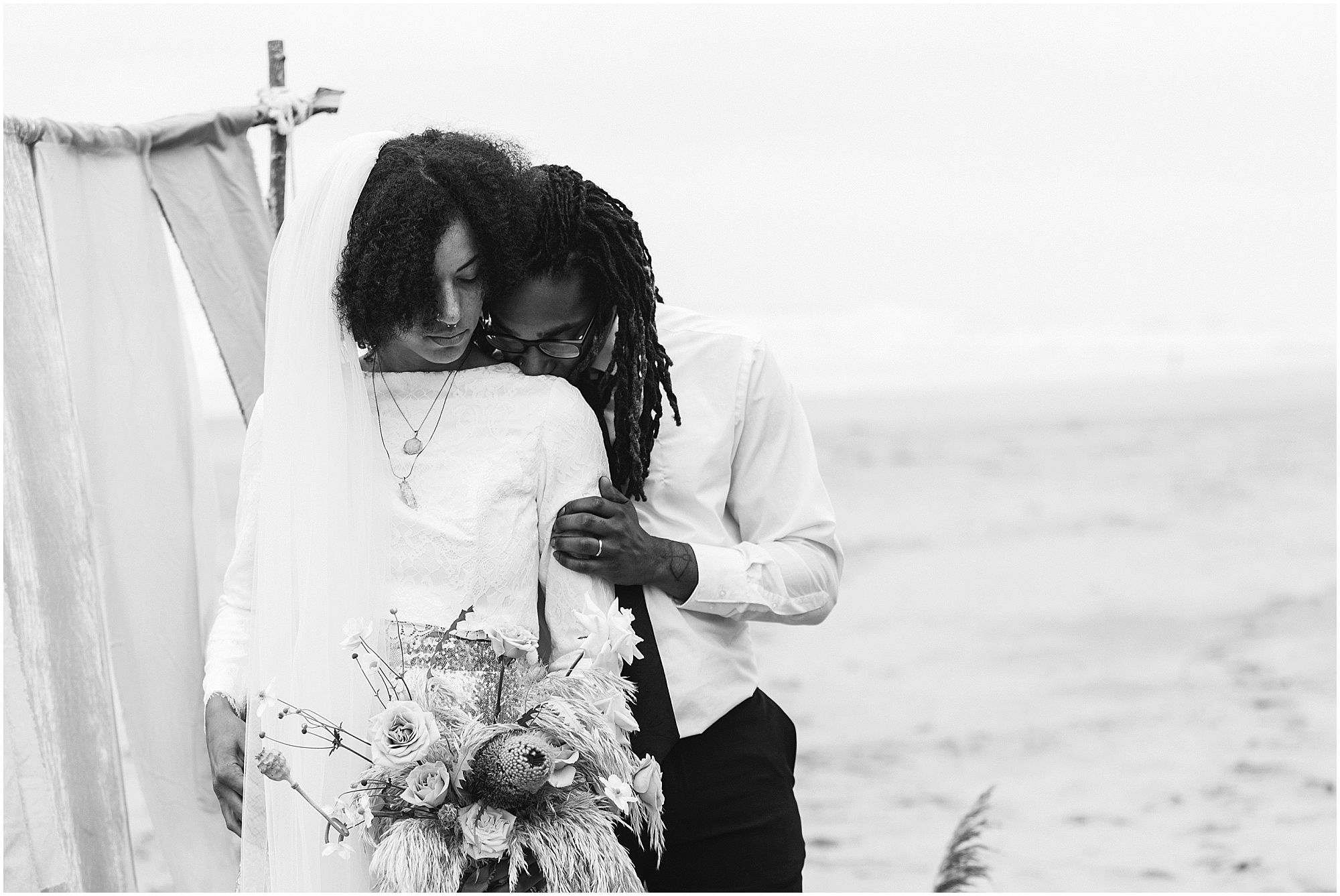 A beautiful black and white image of a groom gently kissing his bride's shoulder in front of their alter on the beach of their Oregon Coast elopement near Lincoln City. | Erica Swantek Photography