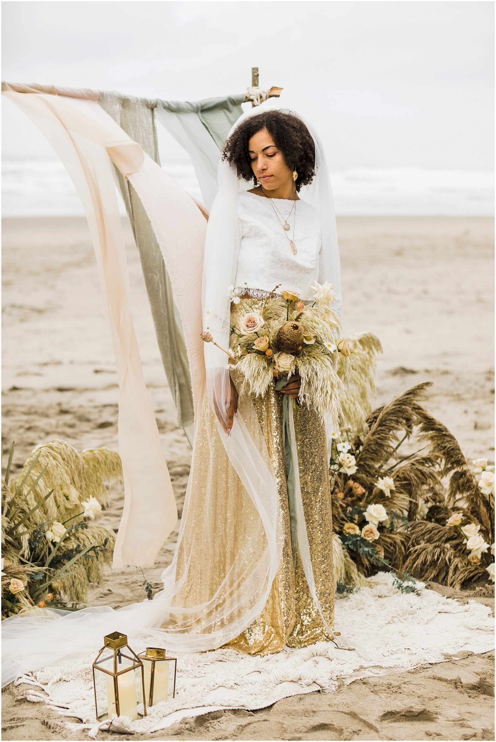 A beautiful black bride, wearing a white long sleeve cropped top, with a gorgeous gold sequined skirt stands on a cream rug in the sand. Behind her is the rustic wood arch with hand dyed silk ribbons in light blush, sage green and cream. Two large floral arrangements made of tall grasses and roses in a neutral color palette frame the arch, with the Pacific Ocean in the backdrop. | Erica Swantek Photography