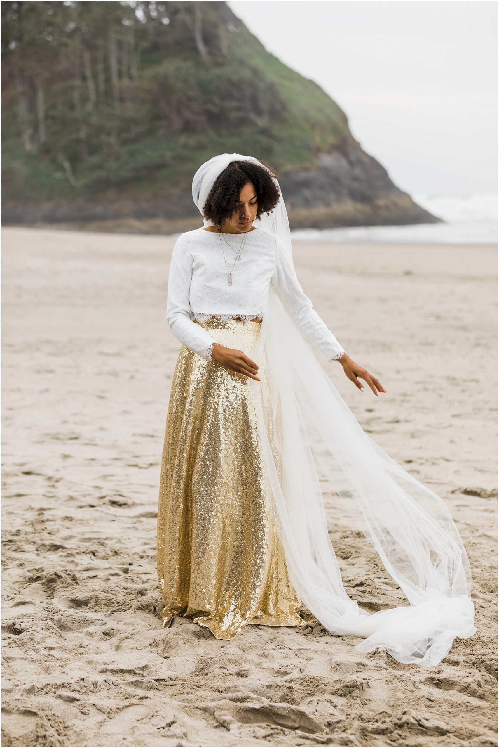 A gorgeous BIPOC bride, wearing a white long sleeve cropped top and gold sequined skirt, plays with her long veil in the sand with Proposal Rock in the backdrop during her Oregon Coast elopement. | Erica Swantek Photography