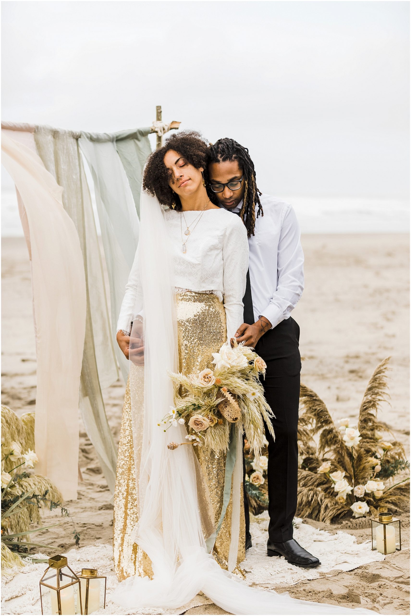 A handsome black groom nuzzles into the shoulder of his beautiful black bride, wearing a white long sleeve cropped top, with a gorgeous gold sequined skirt while standing on a cream rug in the sand. Behind her is the rustic wood arch with hand dyed silk ribbons in light blush, sage green and cream. Two large floral arrangements made of tall grasses and roses in a neutral color palette frame the arch, with the Pacific Ocean in the backdrop. | Erica Swantek Photography